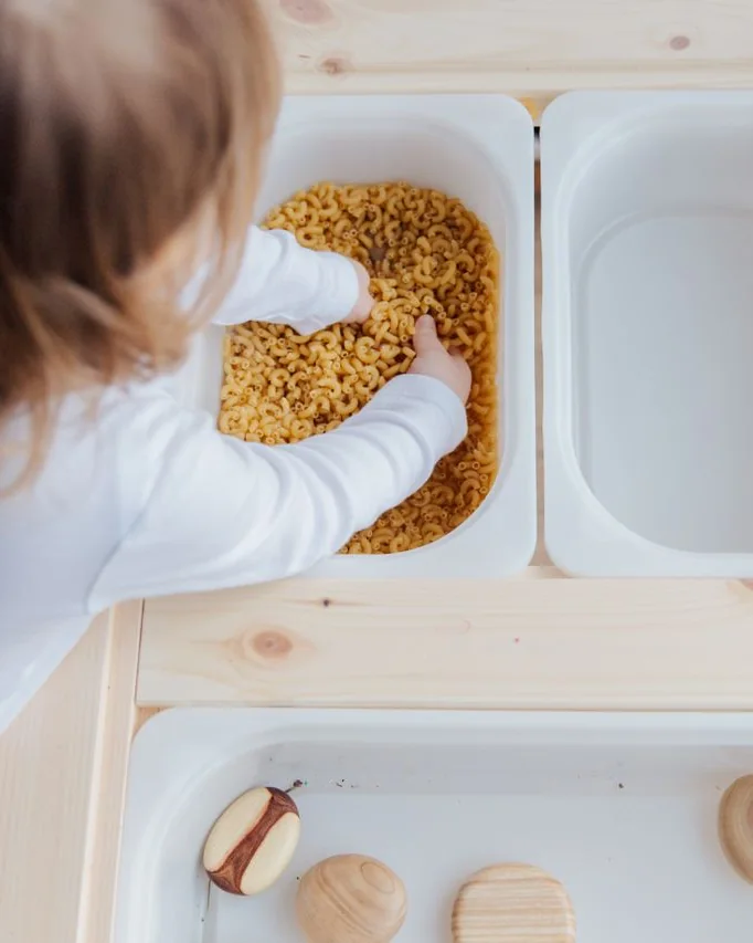 Little hands digging into a tub of dry pasta at a wooden sensory table